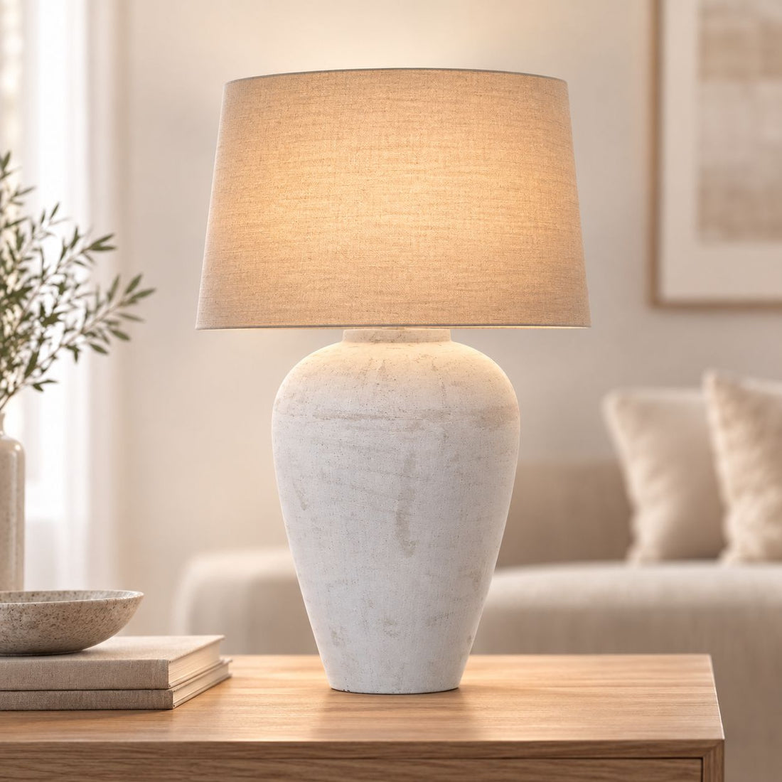 Athena aged stone table lamp lit on a wooden side table beside a sofa