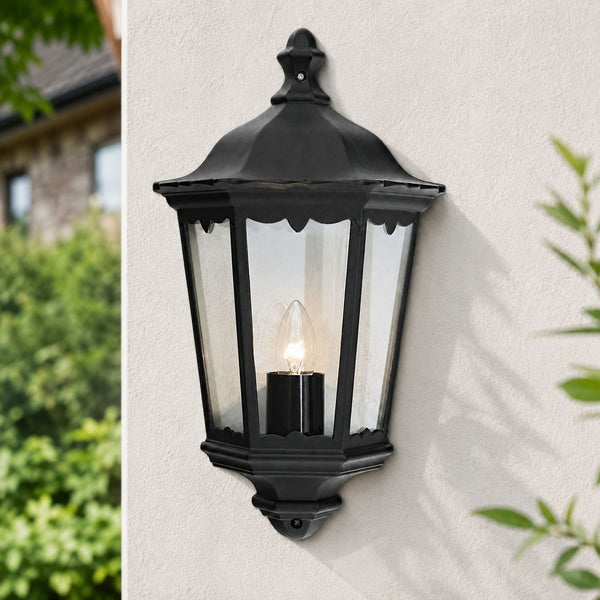 Corben matt black half lantern outdoor wall light lit on a white wall with foliage