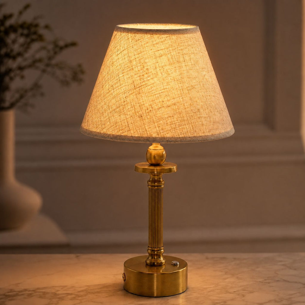 Lira brass cordless table lamp with oatmeal shade lit on a table by a window