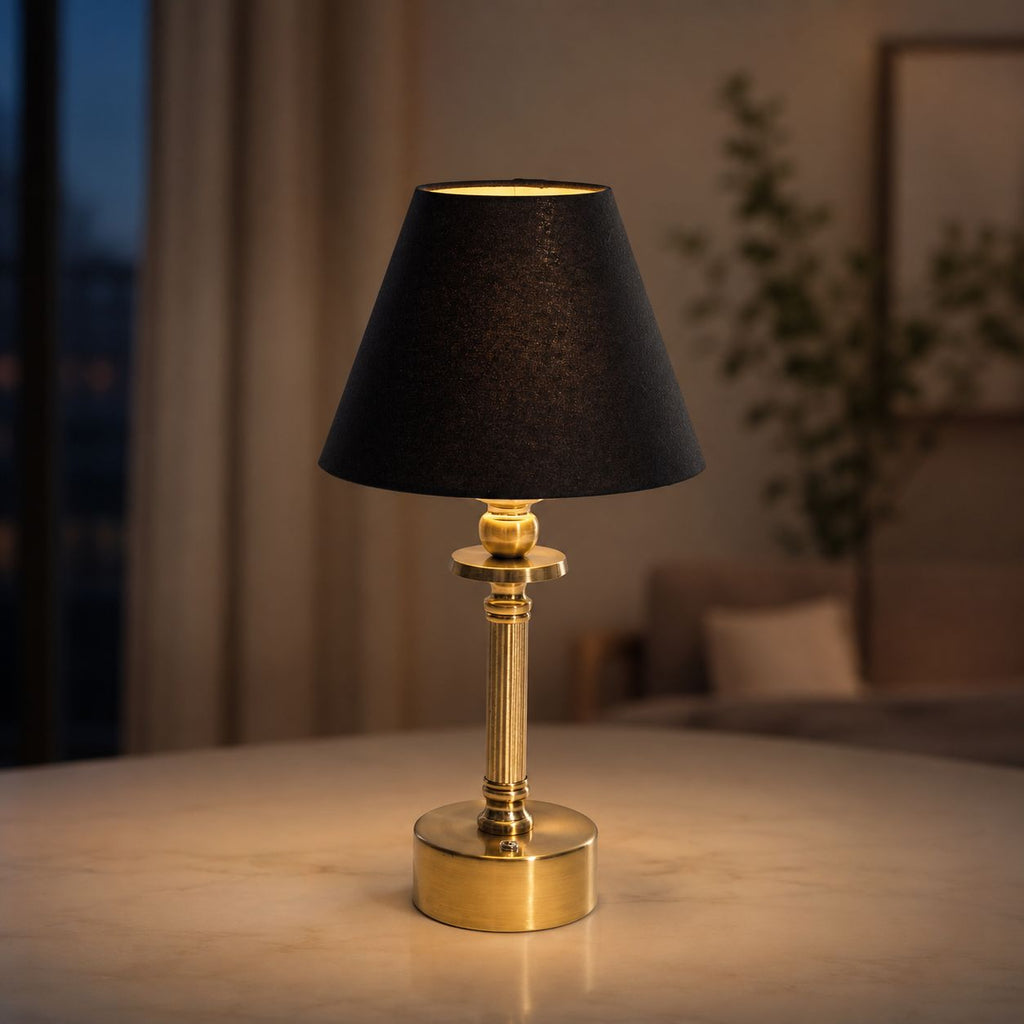 Lira brass cordless table lamp with black shade lit on a table by a window