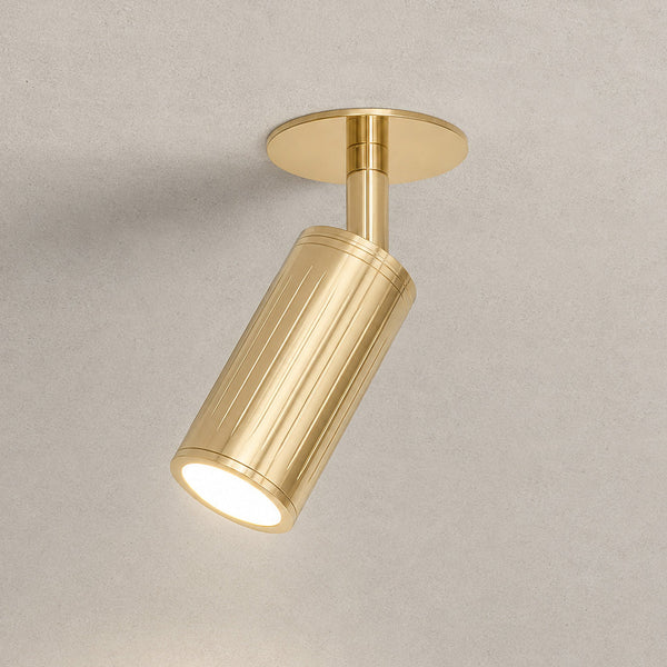 A modern cylindrical track spotlight in a brushed gold finish, shown illuminated against a textured light gray ceiling.