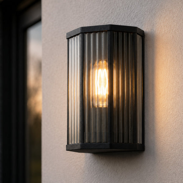 Half lantern outdoor wall light in matt black with ribbed glass, lit at dusk