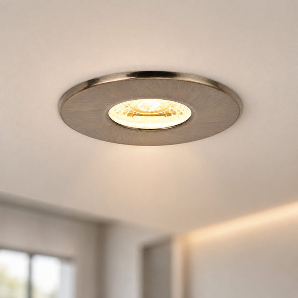 A round recessed ceiling light fixture with a brushed silver metal frame and a warm yellow light glowing from within, installed on a white ceiling in a modern room.