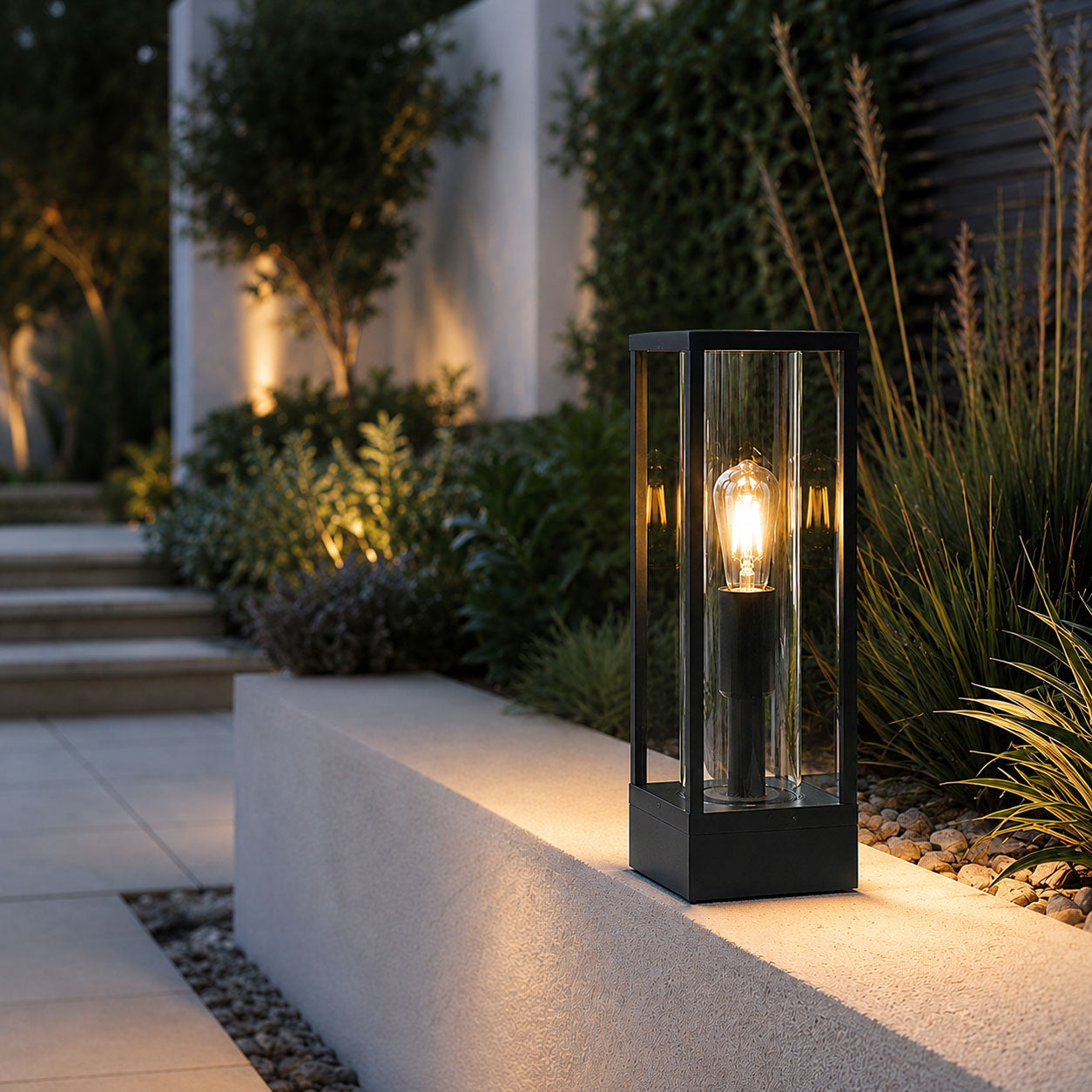 Ted black box post lantern lit at dusk on a patio step beside ornamental grasses