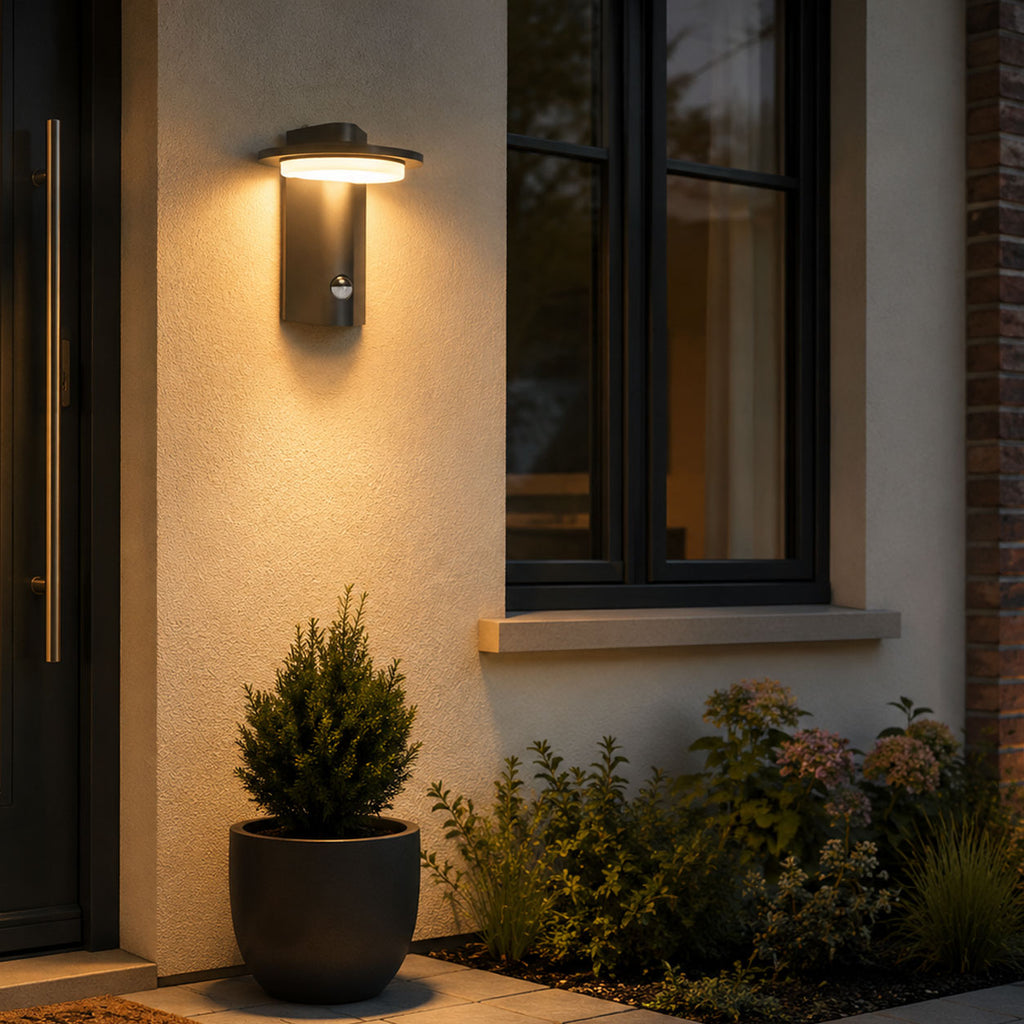 Mayson dark grey LED sensor wall light lit on a rendered porch wall at dusk